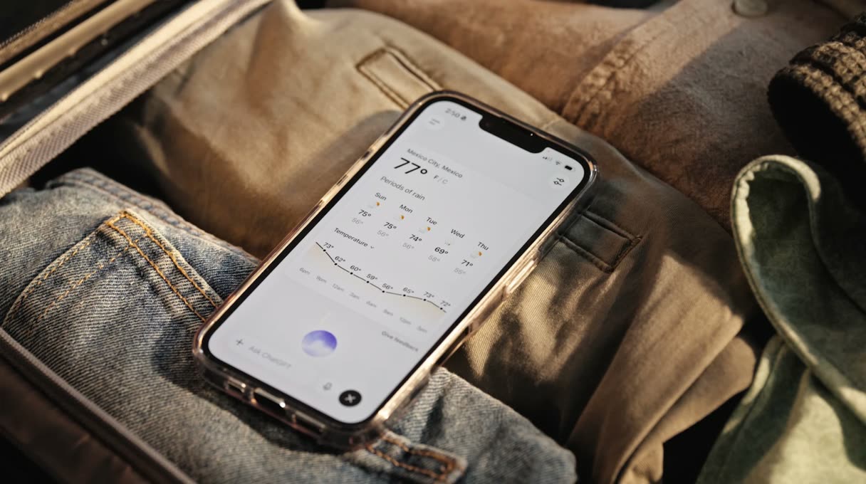 Smartphone mit geöffnetem ChatGPT Voice Mode und Wettervorhersage, liegend auf Kleidung in einem gepackten Koffer